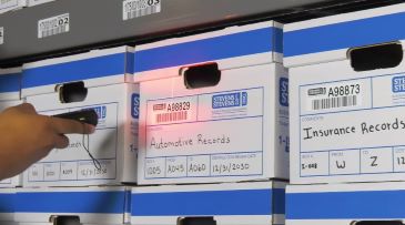 A person is pointing a handheld scanner at the bar code on a document archive box on a shelf.